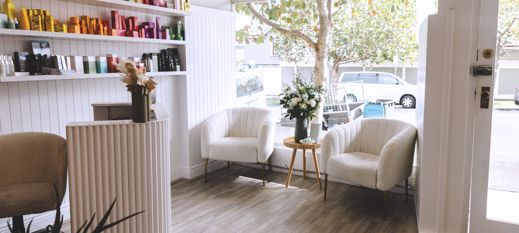 Makeup & Beauty Lounge salon reception in Moonee Ponds featuring two white chairs, product display shelf with beauty essentials, and a modern reception desk – welcoming clients for lashes, brows, tanning, waxing, and body sculpting services.