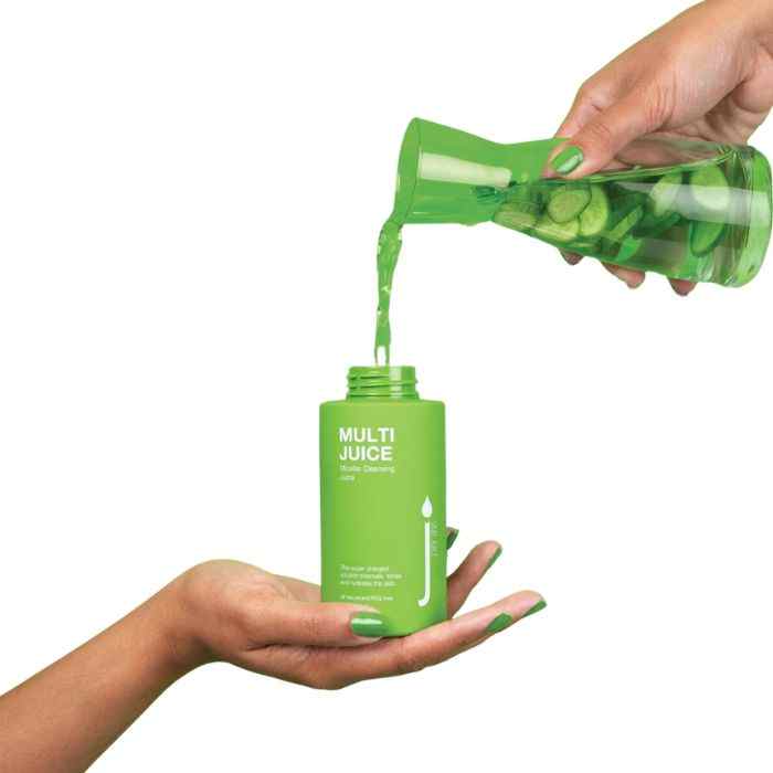 Pouring Multi Juice Micellar Water Cleansing Skin Drink from a green bottle into a matching green container with fresh cucumber slices visible in the liquid.